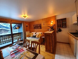 A cozy room with a wooden wall, a sofa, a dining table with magazines, and a kitchen area with a sink and cabinets.