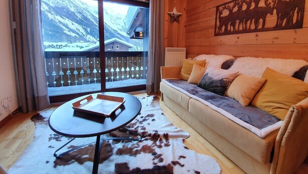 Une pièce chaleureuse avec un canapé, une table sur laquelle repose un livre, un tapis en peau de vache et une vue sur les montagnes, visible à travers de grandes fenêtres.