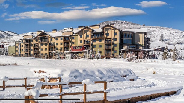 A snow-covered mountain resort with multi-story buildings, ski lifts, and a wooden fence in the foreground.
