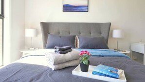 A neatly made bed with a grey headboard, a bedside table with a lamp, a small potted plant, and a tray with books and folded towels.