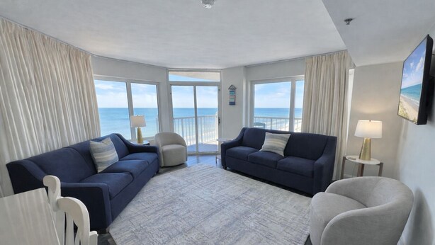 Ein geräumiges Wohnzimmer mit großem Fenster und Meerblick, zwei dunkelblaue Sofas, ein heller Teppich und ein kleiner runder Couchtisch mit einer Lampe.