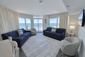 A spacious living room with a large window offering a view of the ocean, two dark blue sofas, a light-colored rug, and a small round table with a lamp. - Seawatch Resort 2 King 2 bath Oceanfront (Myrtle Beach)