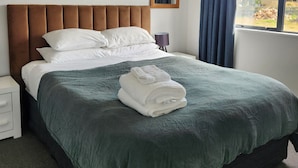 A neatly made bed with a green bedspread and white pillows, a bedside table with a lamp, and a window with curtains. - TWIZEL - House near the lake (Twizel)