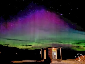 A small cabin with a glass door and windows, surrounded by a natural landscape under a night sky with aurora borealis. - TWIZEL - House near the lake (Twizel)