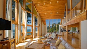 A spacious living room with a large wooden ceiling, multiple windows, a sofa, and a television. - Gorgeous Oceanview! by OceanviewHotTubs Tesla Charging Oceanfront! Shelter Cove (Whitethorn)