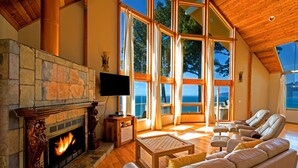 A spacious living room with a stone fireplace, leather sofas, and large windows offering a view of the outdoors. - Gorgeous Oceanview! by OceanviewHotTubs Tesla Charging Oceanfront! Shelter Cove (Whitethorn)