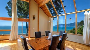 A dining area with a wooden table and chairs, large windows with a view of the sea and trees, and a wooden ceiling. - Gorgeous Oceanview! by OceanviewHotTubs Tesla Charging Oceanfront! Shelter Cove (Whitethorn)