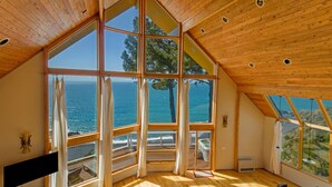 A spacious room with large windows offering ocean views, wooden ceiling, and a cozy seating area. - Gorgeous Oceanview! by OceanviewHotTubs Tesla Charging Oceanfront! Shelter Cove (Whitethorn)