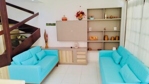Una sala de estar con dos sofás azules, una escalera de madera, un televisor montado en la pared y un estante con adornos.
