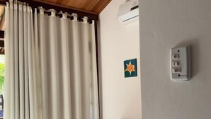 Un dormitorio con cama, cortinas blancas, unidad de aire acondicionado y toma de corriente en la pared.