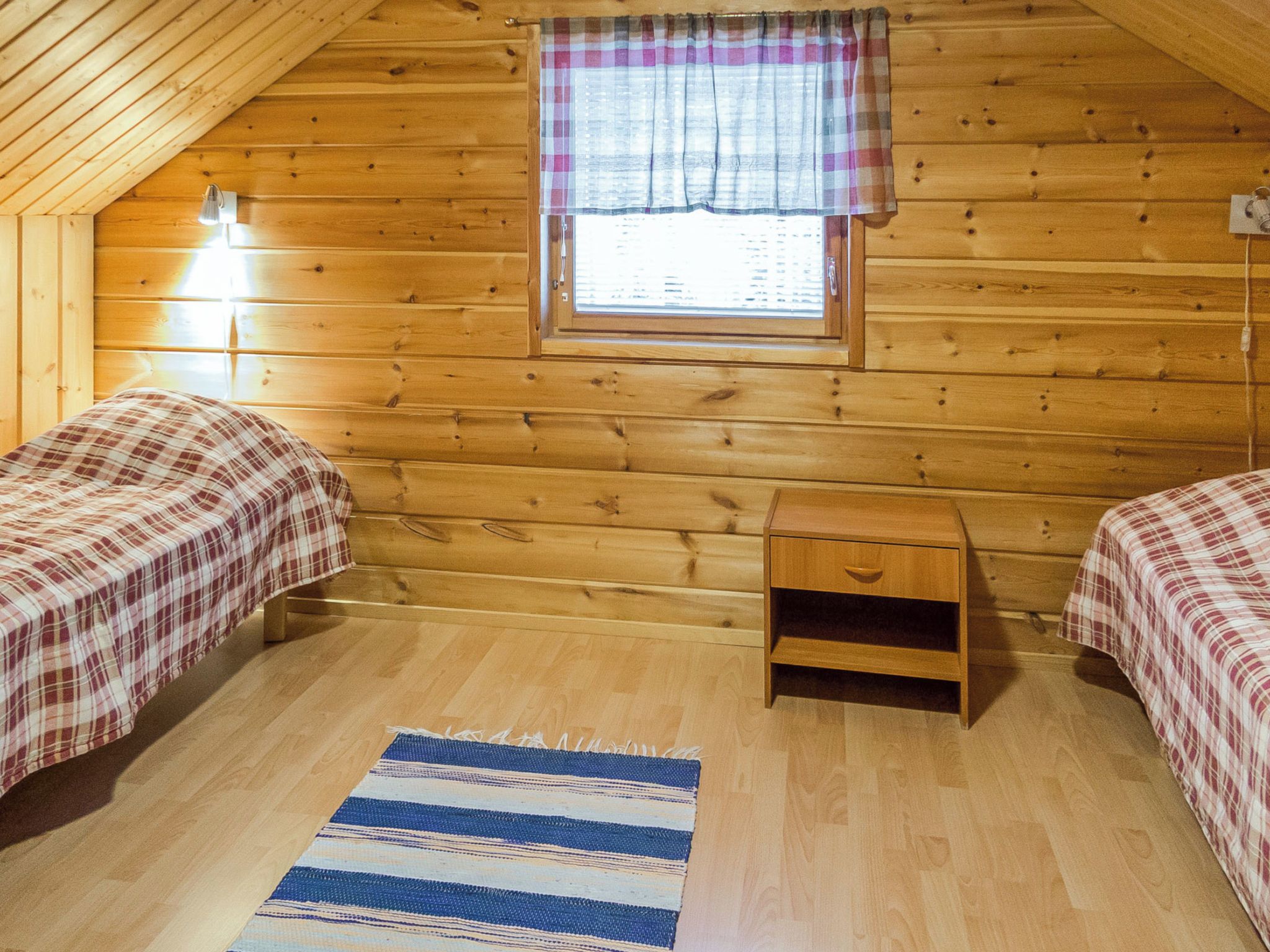 A cozy cabin room with two beds, a small wooden nightstand, a window with a plaid curtain, and a striped rug on the wooden floor.
