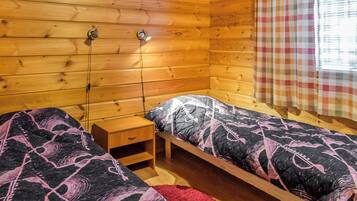 A wooden cabin room with two single beds, a nightstand, and a window with a checkered curtain.