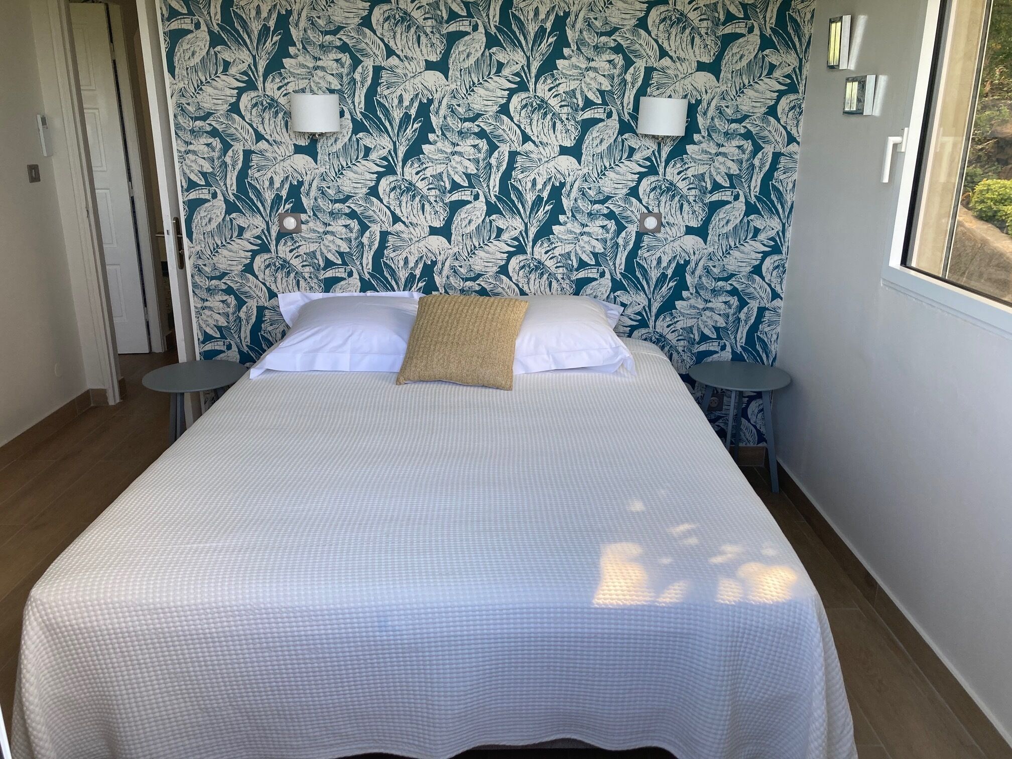 A neatly made bed with a white bedspread and two pillows, a small round table beside it, and a wall with a leafy pattern.