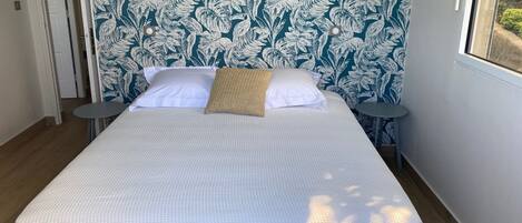A neatly made bed with a white bedspread and two pillows, a small round table beside it, and a wall with a leafy pattern.