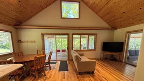 A spacious room with wooden floors, a dining table with chairs, a sofa, a television, and large windows offering views of greenery.