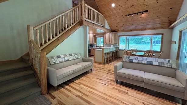 A spacious living area with a wooden staircase, two sofas, a kitchen, and large windows.