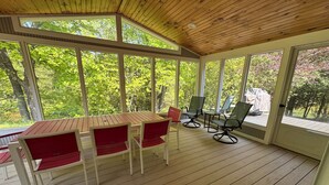 A covered outdoor area with a wooden table and chairs, large windows, and a view of trees.