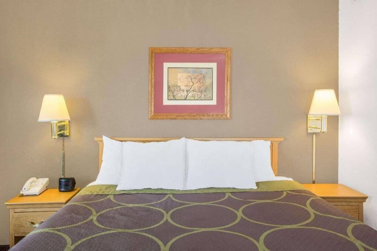 A hotel room with a bed, two bedside lamps, a framed picture on the wall, and a nightstand with a phone and a clock.