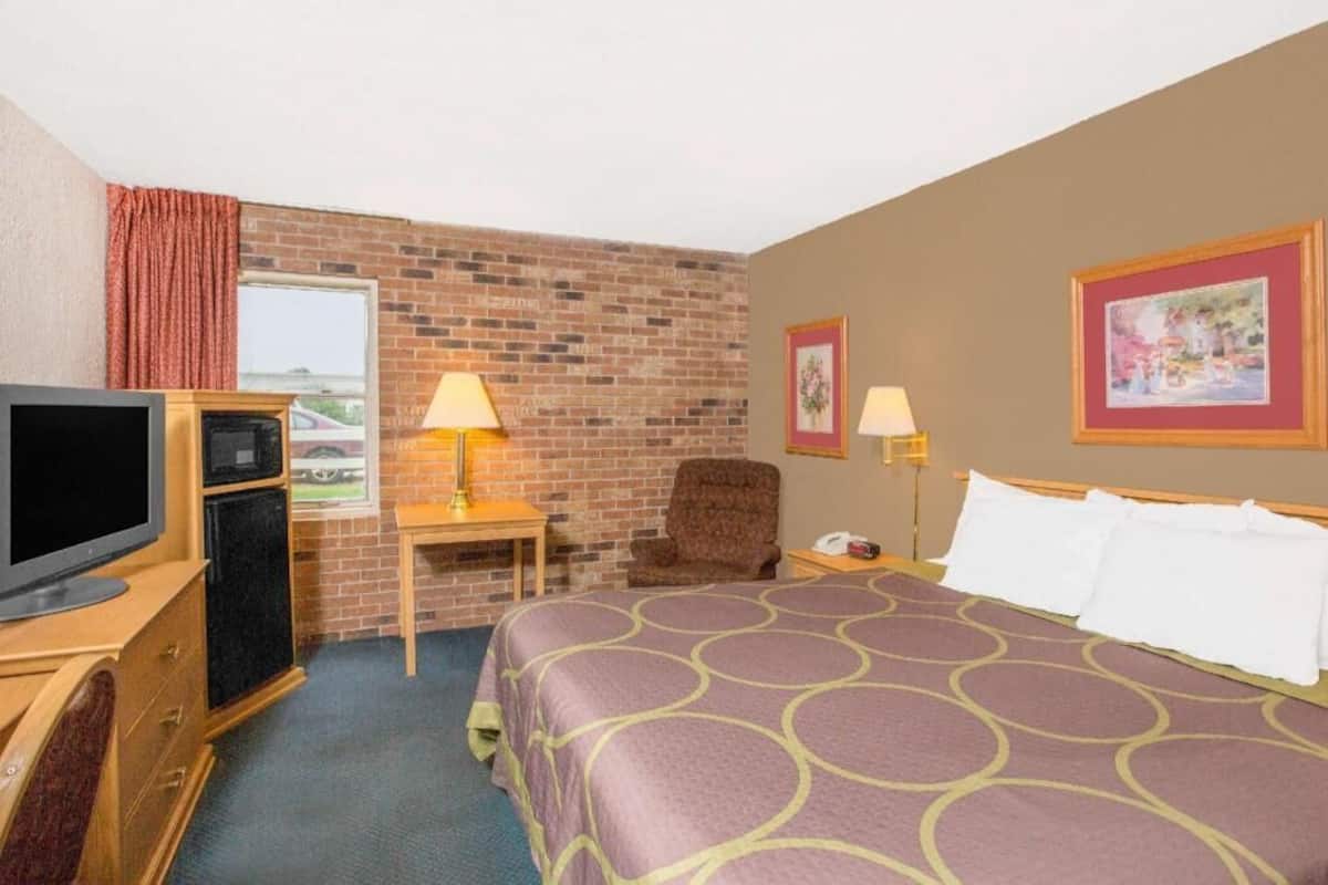 A hotel room with a brick wall, a bed with a patterned bedspread, a television, a desk with a lamp, and a chair.