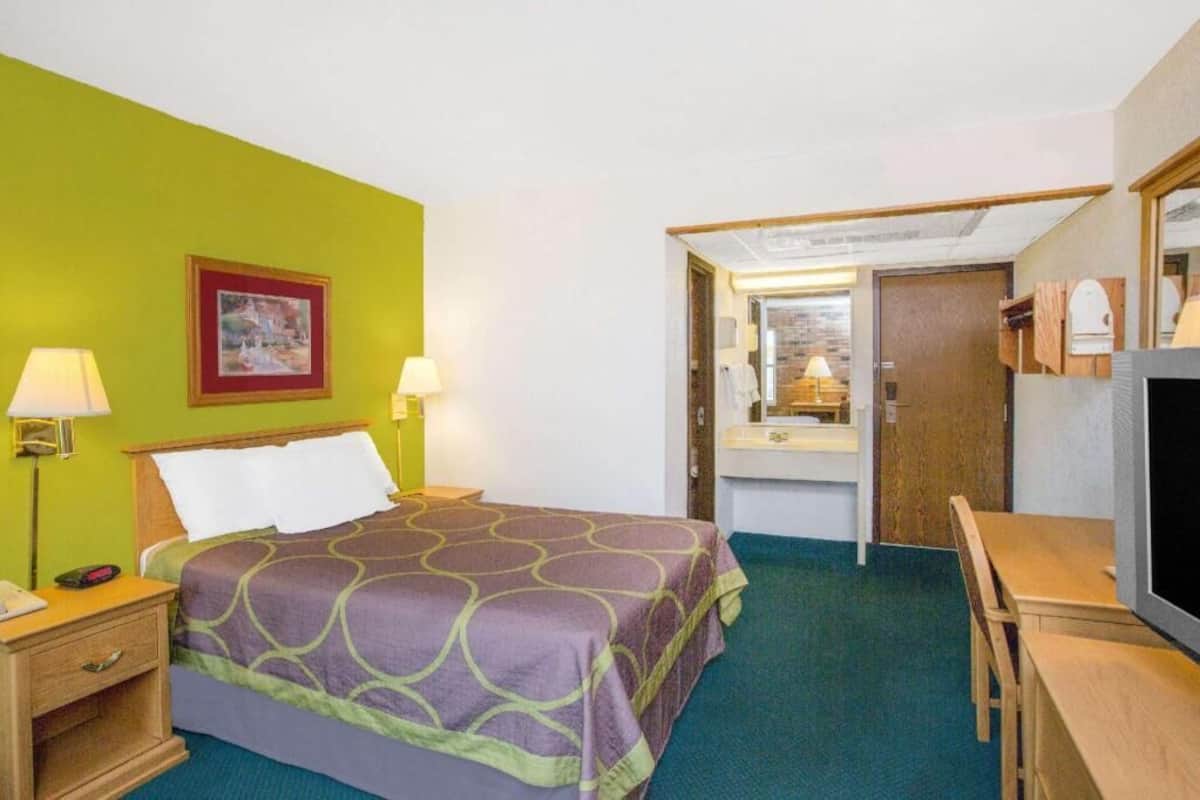 A hotel room with a bed, bedside lamps, a desk, a chair, a mirror, and a bathroom visible through an open door.
