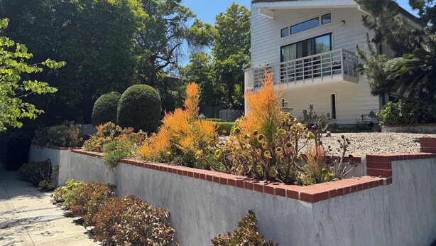 A modern two-story house with a balcony, surrounded by a garden with various plants and a concrete retaining wall. - Large luxury studio with view in of golf course , (Santa Monica)