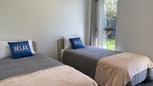 A hotel room with two beds, each with a grey and beige blanket, and a blue pillow with the word "RELAX." - By The Harbour (Tauranga)
