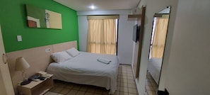 A hotel room with a bed, a bedside table with a lamp, a mirror, a TV, and a window with curtains. - Flat Number One Ferreira Hospdagens (São Luís)