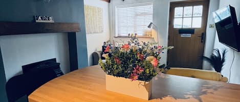 A room with a wooden table, a flower vase, a chair, a desk, a lamp, a television, and a door with a window.