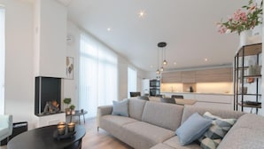 A modern living room with a sofa, coffee table, and fireplace. There is a kitchen in the background with a dining area.