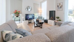 A modern living room with a large grey sofa, a flat-screen TV, a fireplace, and a large window.
