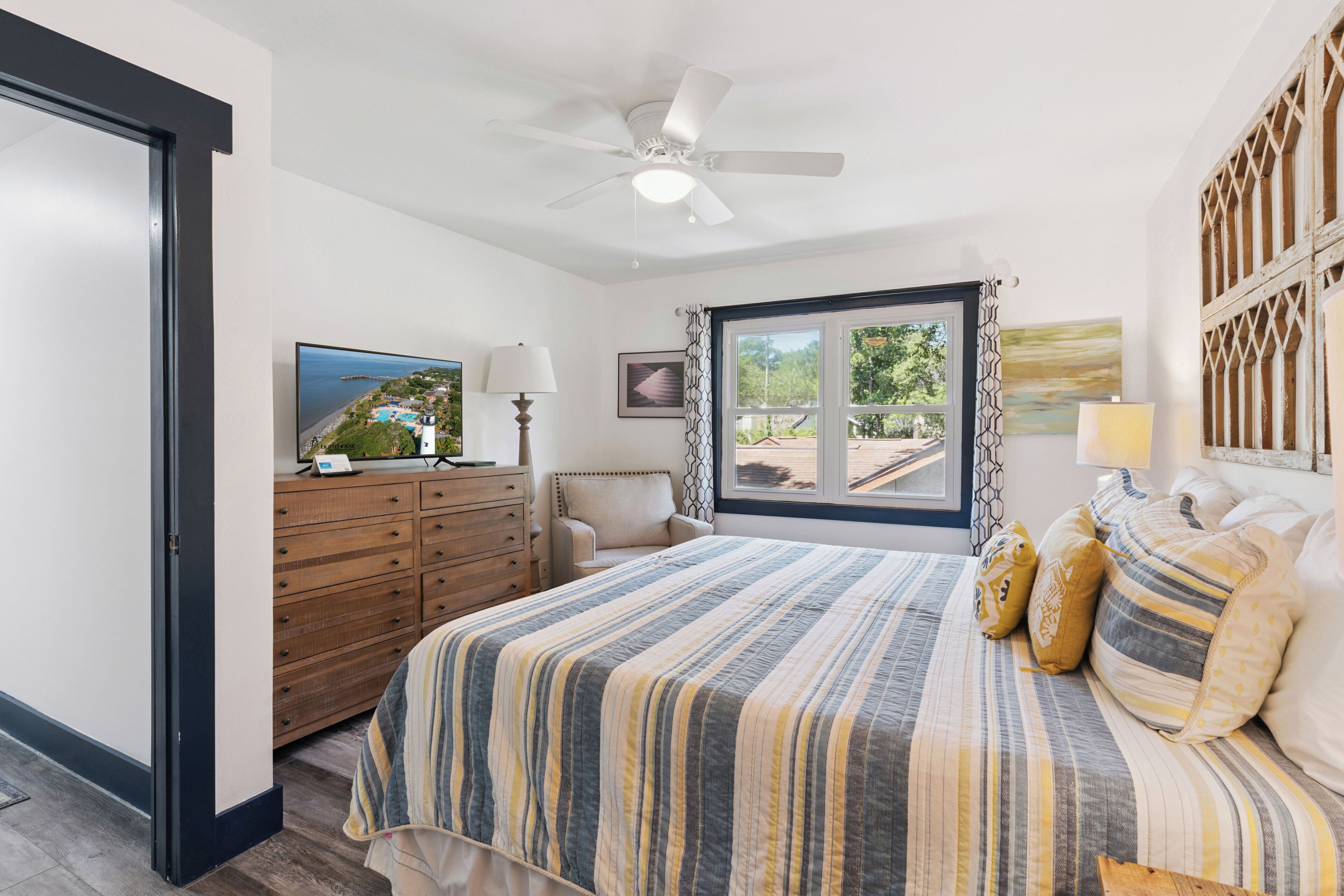 A bedroom with a bed, a ceiling fan, a window with curtains, a dresser, a chair, and a television.