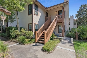 A two-story building with a wooden staircase leading to a balcony, surrounded by greenery and a clear sky.