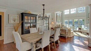A dining room with a wooden table and chairs, a chandelier, and large windows. - Waterfront Villa near Hamptons with pool, hot tub & boat dock (Center Moriches)