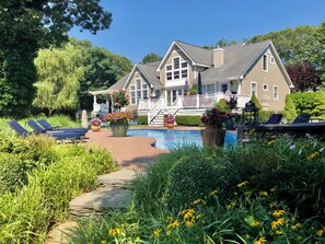 A two-story house with a swimming pool, sun loungers, and a garden with flowers.