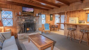 Un interior acogedor de cabaña con chimenea de piedra, muebles de madera, televisor y una zona de cocina.