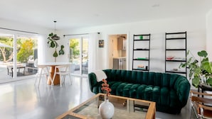 A modern living room with a green velvet sofa, a glass coffee table, and a dining area with white chairs.