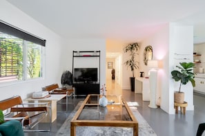 Un salon moderne avec une table basse en verre, des chaises en bois, un téléviseur à écran plat et une grande fenêtre équipée de stores.