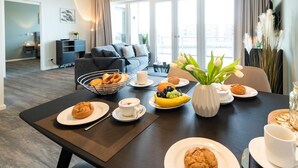 Een moderne woonkamer met een eethoek, een bank, een salontafel met ontbijtartikelen en een groot raam met gordijnen.