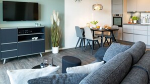 Een moderne woonkamer met een flatscreen televisie, een grijze bank, een eettafel, stoelen en een keuken op de achtergrond.