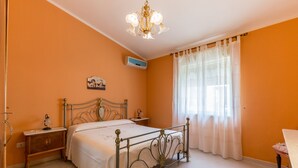 Une chambre avec un lit en métal, une table de chevet, une commode, un climatiseur fixé au mur et une fenêtre avec des rideaux légers.