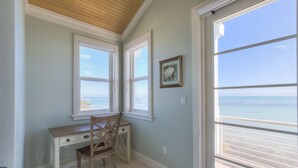 Una habitación con escritorio y silla, ventanas amplias con vistas al océano, y un cuadro en la pared.