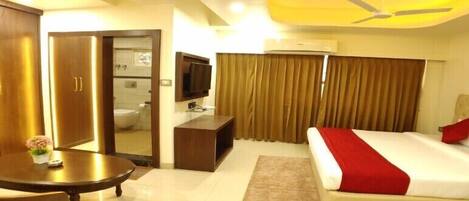 A hotel room with a bed, a desk, a chair, a television, a ceiling fan, and a bathroom visible through an open door.