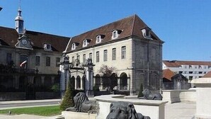 A courtyard with a fountain featuring lion statues, surrounded by a building with a clock tower and arched windows. - STUDIO LILOU (Lons-le-Saunier)