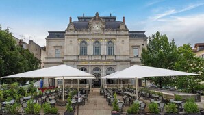 A grand cafe with outdoor seating, a clock, and a theater sign. - STUDIO LILOU (Lons-le-Saunier)