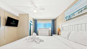 Una camera d'albergo con un letto, una ventola a soffitto, una televisione, un quadro e vista sulla spiaggia.