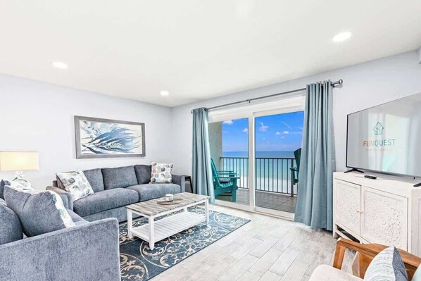 A modern living room with a grey sofa, a coffee table, and a flat-screen TV on a white cabinet. A large window offers a view of the sea.