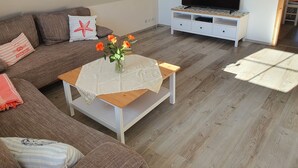 A cozy living room with a brown sofa, a white coffee table with a vase of flowers, and a flat-screen TV on a white stand. - Schöne Ferienwohnung in Ruhiger Wohnsiedlung mit Unverbautem Feldblick (Wankendorf)
