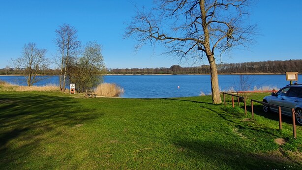 A grassy area with a lake, a tree, and a car parked near a railing. - Schöne Ferienwohnung in Ruhiger Wohnsiedlung mit Unverbautem Feldblick (Wankendorf)