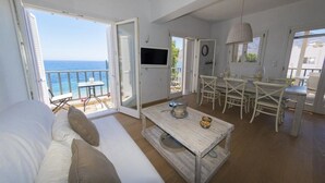 Ein geräumiger Wohnbereich mit Balkon und Meerblick, ein Esstisch sowie ein an der Wand befestigter Fernseher.