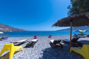 Ein Kiesstrand mit Liegestühlen und Sonnenschirmen, ein klares blaues Meer und eine gebirgige Kulisse.